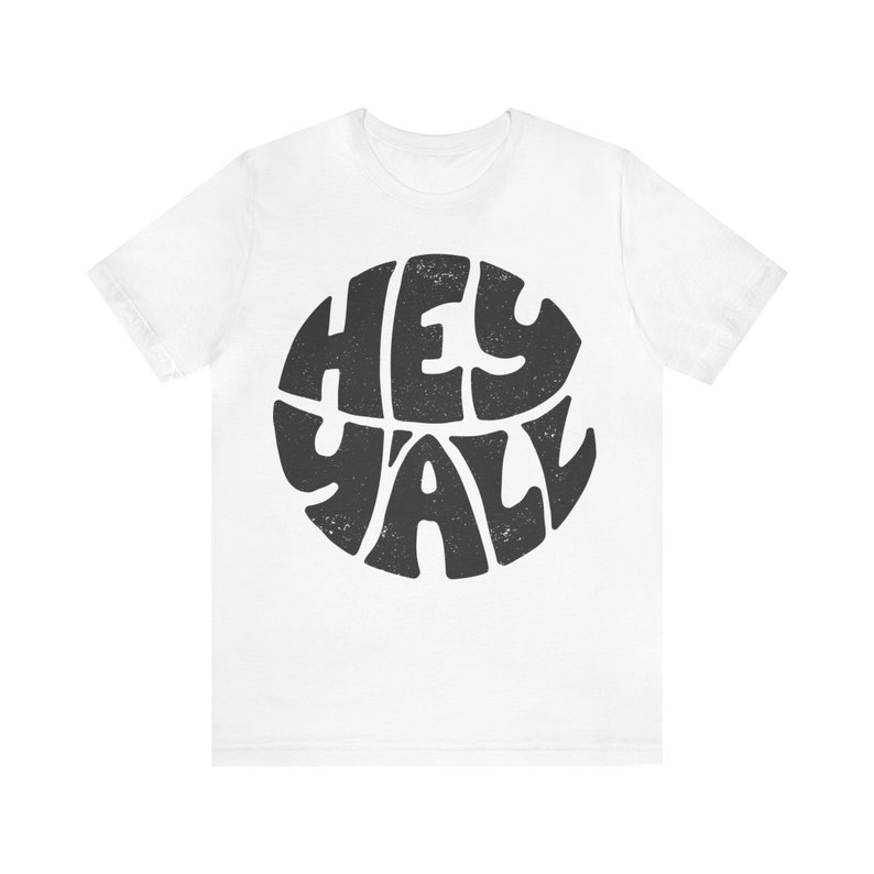 Pu&ograve; includere: Maglietta bianca con un grafico circolare nero che dice "Hey Y'all" in un font retr&ograve;.