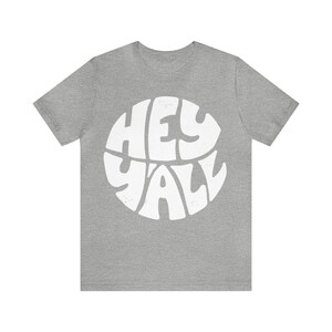 Pu&ograve; includere: Una t-shirt color grigio melange con una grafica bianca del testo "Hey Y'all" in un design circolare.