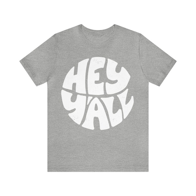 Pu&ograve; includere: Una t-shirt color grigio melange con una grafica bianca del testo "Hey Y'all" in un design circolare.