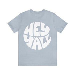 Pu&ograve; includere: Una t-shirt blu chiaro con una grafica circolare bianca che dice "Hey Y'all" in un font retr&ograve;.
