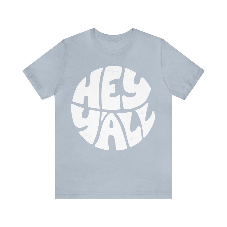 Pu&ograve; includere: Una t-shirt blu chiaro con una grafica circolare bianca che dice "Hey Y'all" in un font retr&ograve;.
