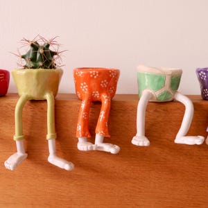 May include: A selection of quirky ceramic plant pots, each with a distinctive design. One pot contains a cactus. The pots are in various colours, including yellow, orange, and green, and feature legs and feet, adding a playful touch to home decor.