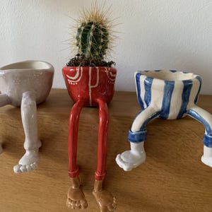 Puede incluir: Tres macetas de cer&aacute;mica con patas. Una es rosa claro, otra roja con detalles blancos y la tercera a rayas azules y blancas. La maceta roja contiene un cactus. Las macetas est&aacute;n sobre una superficie de madera.