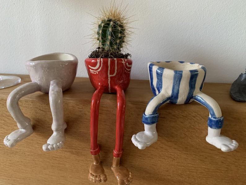 Puede incluir: Tres macetas de cer&aacute;mica con patas. Una es rosa claro, otra roja con detalles blancos y la tercera a rayas azules y blancas. La maceta roja contiene un cactus. Las macetas est&aacute;n sobre una superficie de madera.