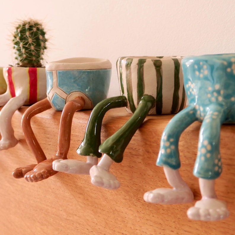 Puede incluir: Una fila de macetas de cer&aacute;mica caprichosas con patas. Las macetas est&aacute;n pintadas en varios colores y dise&ntilde;os, incluyendo rayas y lunares. Cada maceta tiene un dise&ntilde;o &uacute;nico, con patas que cuelgan del borde de una estanter&iacute;a de madera.