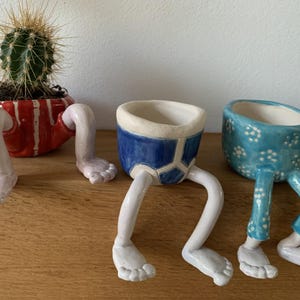 Puede incluir: Tres macetas de cer&aacute;mica caprichosas con patas. Una maceta a rayas rojas y blancas contiene un cactus. Tambi&eacute;n se muestra una maceta azul con ribete blanco y una maceta azul con lunares blancos.