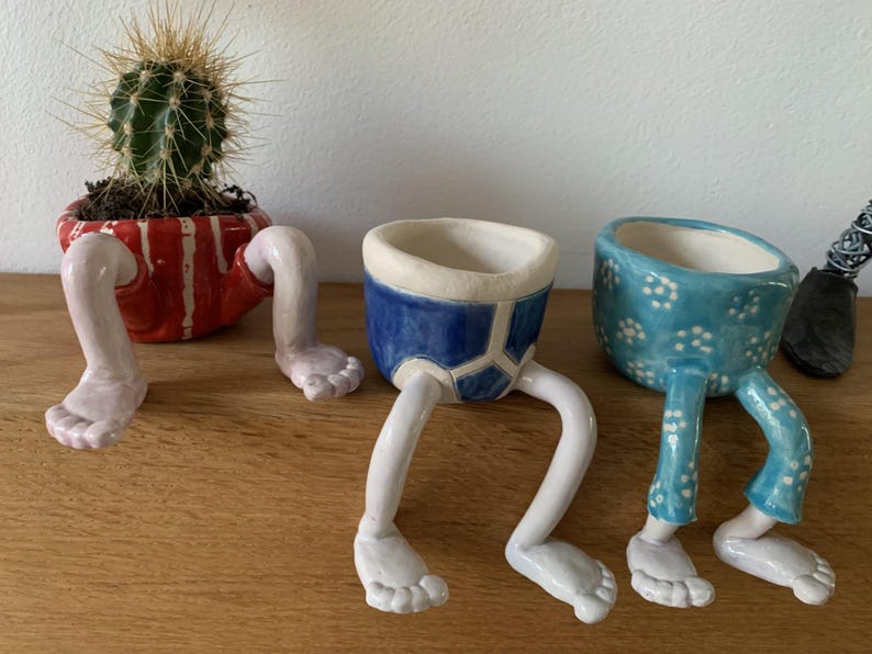 Puede incluir: Tres macetas de cer&aacute;mica caprichosas con patas. Una maceta a rayas rojas y blancas contiene un cactus. Tambi&eacute;n se muestra una maceta azul con ribete blanco y una maceta azul con lunares blancos.