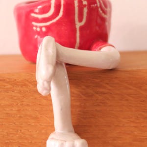Puede incluir: Una maceta de cer&aacute;mica caprichosa con forma de pantal&oacute;n corto rojo con detalles blancos y piernas blancas adjuntas. La maceta est&aacute; dise&ntilde;ada para colocarse sobre una superficie, con las piernas extendidas hacia abajo. El dise&ntilde;o general es l&uacute;dico y decorativo.