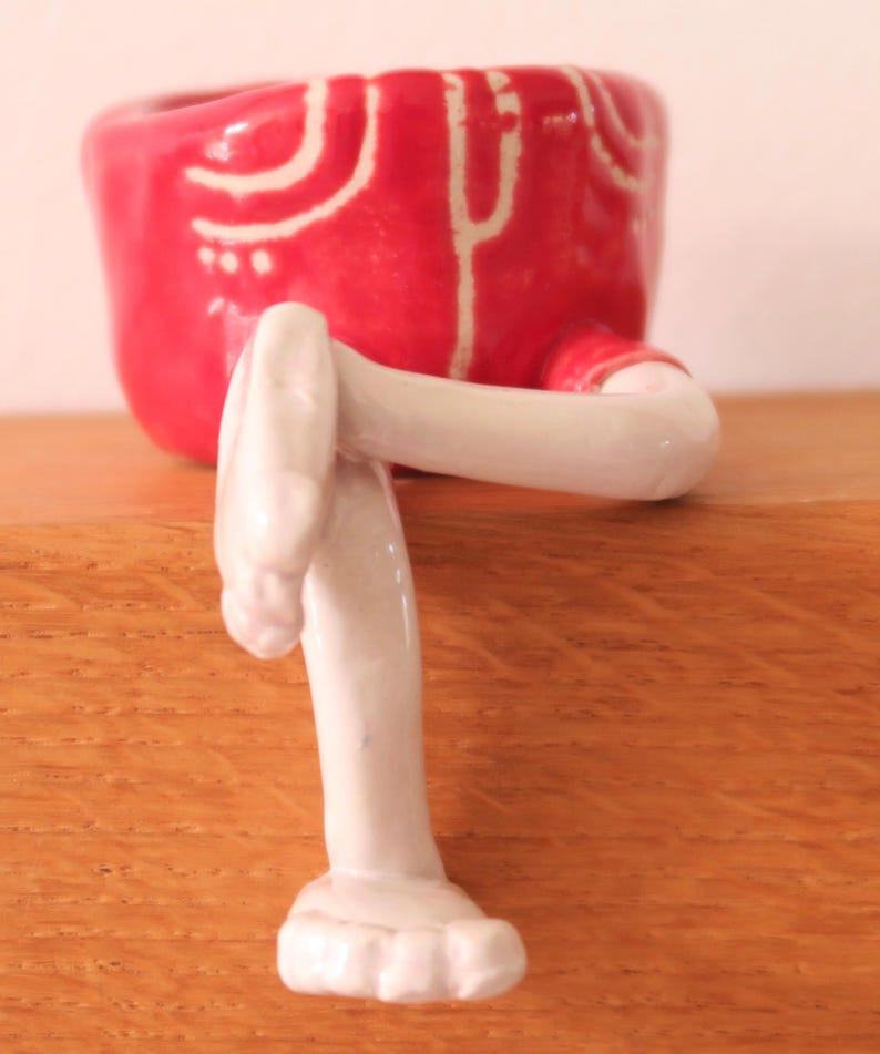Puede incluir: Una maceta de cer&aacute;mica caprichosa con forma de pantal&oacute;n corto rojo con detalles blancos y piernas blancas adjuntas. La maceta est&aacute; dise&ntilde;ada para colocarse sobre una superficie, con las piernas extendidas hacia abajo. El dise&ntilde;o general es l&uacute;dico y decorativo.