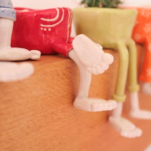 Puede incluir: Primer plano de varias macetas de cer&aacute;mica coloridas con piernas y pies. La maceta roja tiene detalles en blanco, mientras que las otras son verdes y naranjas. Las macetas est&aacute;n dise&ntilde;adas para parecer que est&aacute;n sentadas.