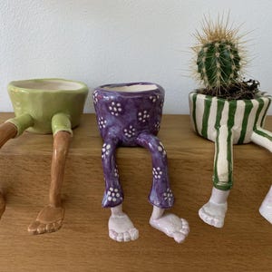 Puede incluir: Tres macetas de cer&aacute;mica caprichosas con patas. Una es verde con patas marrones, otra es morada con lunares blancos, y la tercera es blanca con rayas verdes, que sostiene un cactus. Estas macetas &uacute;nicas a&ntilde;aden un toque l&uacute;dico a cualquier espacio.
