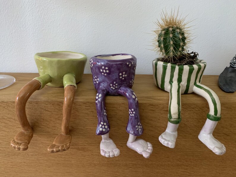 Puede incluir: Tres macetas de cer&aacute;mica caprichosas con patas. Una es verde con patas marrones, otra es morada con lunares blancos, y la tercera es blanca con rayas verdes, que sostiene un cactus. Estas macetas &uacute;nicas a&ntilde;aden un toque l&uacute;dico a cualquier espacio.