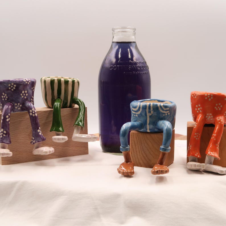 Puede incluir: Una colecci&oacute;n de macetas de cer&aacute;mica caprichosas en varios colores, incluyendo morado, verde, azul y naranja. Cada maceta est&aacute; dise&ntilde;ada con piernas y pies, d&aacute;ndoles una apariencia juguetona y antropom&oacute;rfica. Una botella alta de color morado oscuro se encuentra en el fondo.