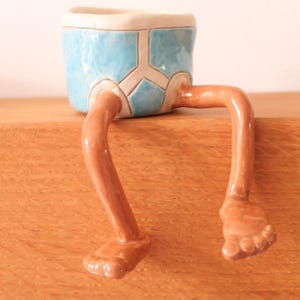 Puede incluir: Una maceta de cer&aacute;mica caprichosa con forma de pantal&oacute;n corto azul con piernas y pies marrones. La maceta descansa sobre una superficie de madera, a&ntilde;adiendo un toque l&uacute;dico a la decoraci&oacute;n del hogar.