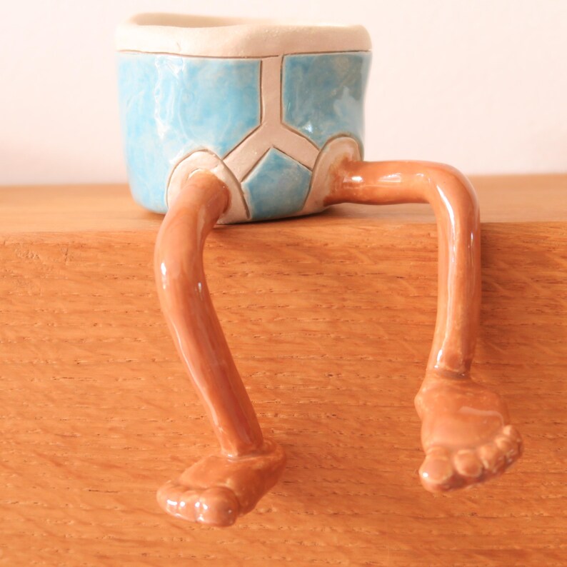 Puede incluir: Una maceta de cer&aacute;mica caprichosa con forma de pantal&oacute;n corto azul con piernas y pies marrones. La maceta descansa sobre una superficie de madera, a&ntilde;adiendo un toque l&uacute;dico a la decoraci&oacute;n del hogar.