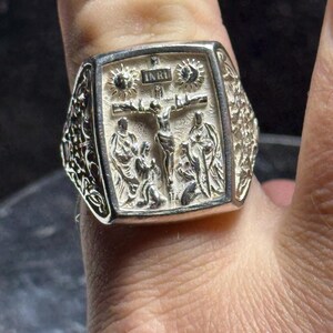 Pode incluir: Um anel de prata com um design detalhado que apresenta um crucifixo e a inscri&ccedil;&atilde;o "INRI". O anel &eacute; usado em um dedo.