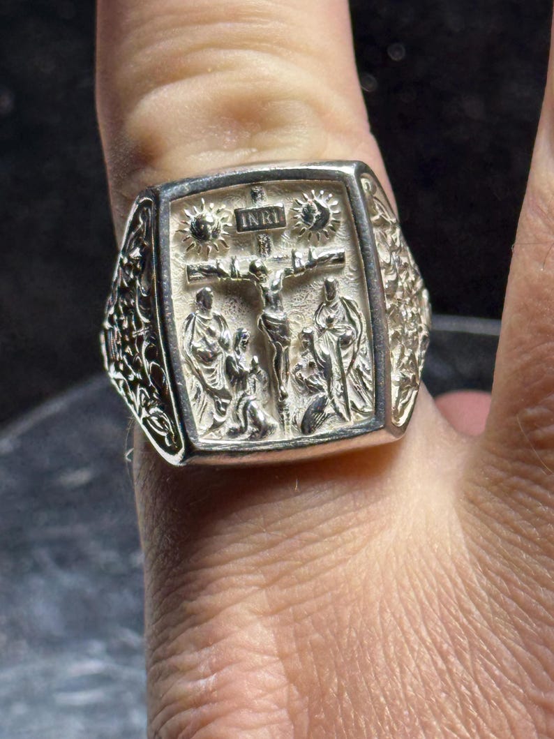 Pode incluir: Um anel de prata com um design detalhado que apresenta um crucifixo e a inscri&ccedil;&atilde;o "INRI". O anel &eacute; usado em um dedo.
