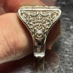 Pode incluir: Um anel de prata com um design intrincado e detalhado. O anel tem uma parte superior de forma quadrada com um padr&atilde;o floral.