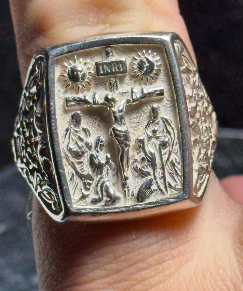 Pode incluir: Um anel de prata com um design detalhado que apresenta um crucifixo e a inscri&ccedil;&atilde;o "INRI". O anel tem uma face quadrada com um design em relevo e detalhes intrincados nas laterais.