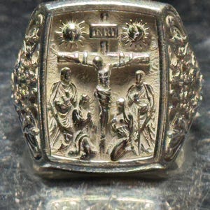 Pode incluir: Um anel de prata com um design detalhado que apresenta uma figura de Jesus Cristo na cruz, rodeado por anjos e um sol radiante. O anel tem uma superf&iacute;cie texturizada e uma borda elevada.