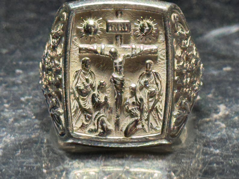 Pode incluir: Um anel de prata com um design detalhado que apresenta uma figura de Jesus Cristo na cruz, rodeado por anjos e um sol radiante. O anel tem uma superf&iacute;cie texturizada e uma borda elevada.