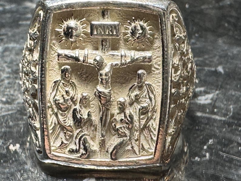 Pode incluir: Um anel de prata com um design detalhado que apresenta um crucifixo e figuras. O anel tem a inscri&ccedil;&atilde;o "INRI" na cruz.