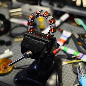 May include: A close-up of a copper-colored coil of wire held in a black metal vise. The coil is wrapped in a clear plastic material and has a circular shape. The vise is on a workbench with various tools and materials scattered around it.