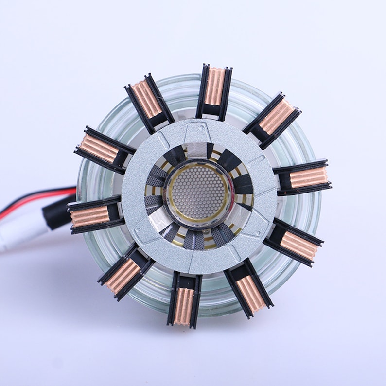May include: A silver and black Iron Man Arc Reactor replica with a clear plastic outer shell and copper-colored accents. The reactor has a circular design with a honeycomb pattern in the center.