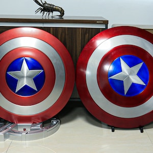 May include: Two red, white, and blue Captain America shields with silver accents. The shields are on stands and are displayed on a wooden surface.