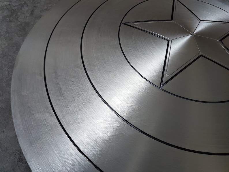 May include: A close-up of a silver Captain America shield with a black star in the center. The shield has a brushed metal finish and concentric circles around the star.