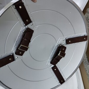 May include: A silver-colored round shield with three brown leather straps attached to the back. The straps have metal buckles.