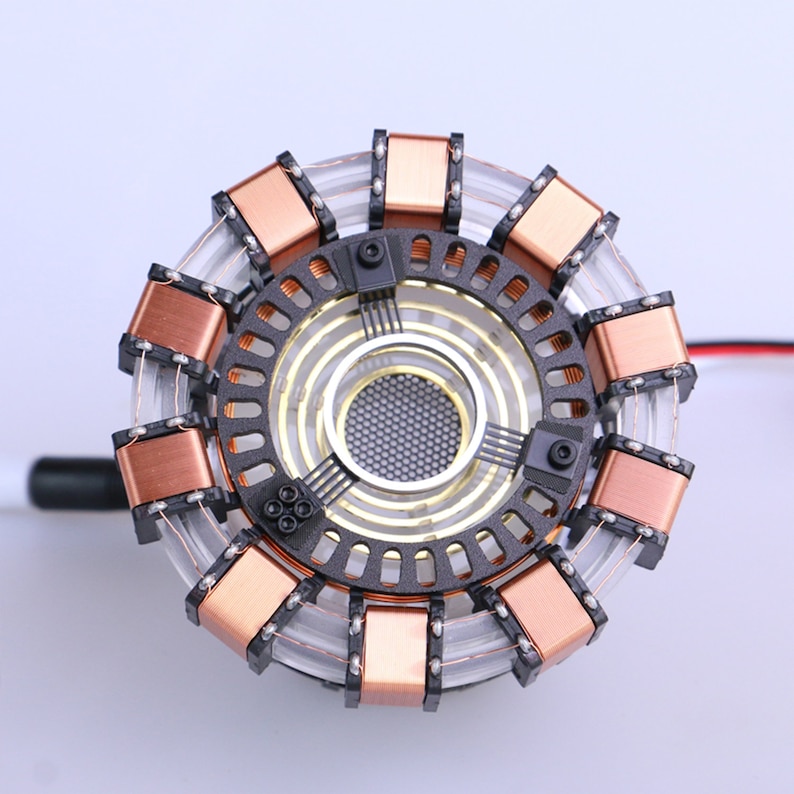 May include: A close-up of a replica of Iron Man's Arc Reactor. The reactor is made of metal and has a copper-colored outer ring with a black center. The center has a circular, silver-colored mesh and a series of black wires.