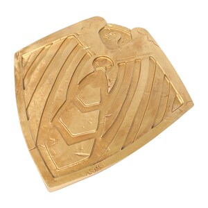 May include: A gold-colored metal shield with a stylized eagle design. The shield has a textured surface and a raised edge.