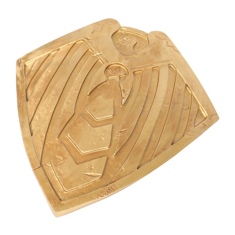 May include: A gold-colored metal shield with a stylized eagle design. The shield has a textured surface and a raised edge.