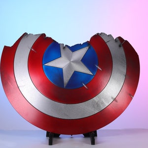 May include: A distressed Captain America shield, featuring a red, white, and blue color scheme. The shield has a star in the center and shows signs of battle damage. The shield is displayed on a black stand.