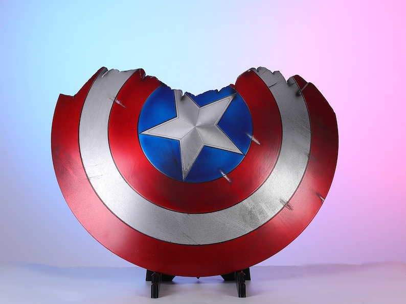 May include: A distressed Captain America shield, featuring a red, white, and blue color scheme. The shield has a star in the center and shows signs of battle damage. The shield is displayed on a black stand.