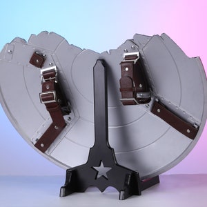 May include: A silver shield with a distressed design, featuring brown leather straps and metal buckles. The shield is displayed on a black stand with a star cutout, set against a gradient background.