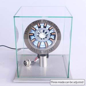 May include: A silver and copper-colored Iron Man Arc Reactor replica with a clear light in the center. The replica is in a glass display case with the text "Proof that Tony Stark has a heart" around the edge.