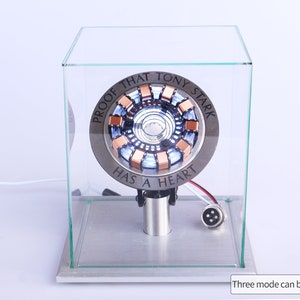 May include: A silver and copper-coloured Iron Man Arc Reactor replica with a clear acrylic display case. The reactor is lit up with blue and white lights. The text "PROOF THAT TONY STARK HAS A HEART" is inscribed around the reactor.