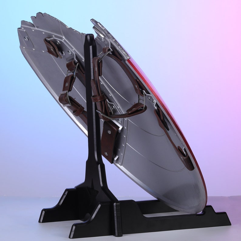 May include: A silver and red shield with brown leather straps, displayed on a black stand. The shield has a star design and is slightly angled, showcasing its metallic finish and details. The stand provides a stable base.