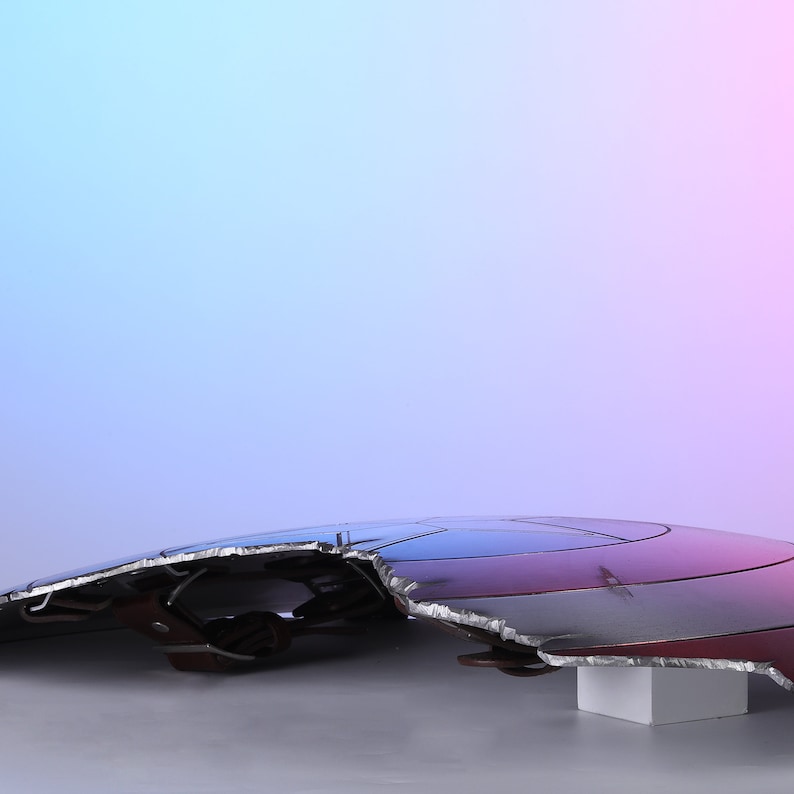 May include: A distressed, metallic shield with a red, white, and blue color scheme. The shield has a jagged edge and is resting on a white block. The background gradient transitions from blue to pink.