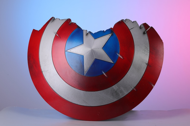 May include: A distressed Captain America shield, featuring a red, white, and blue color scheme. The shield has a star in the center and shows signs of wear and tear, with a portion of the top edge missing. The shield is approximately 24 inches in diameter.
