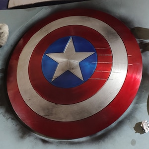 May include: A red, white, and blue Captain America shield with a five-pointed star in the center. The shield has a worn, distressed look.