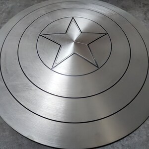 May include: A silver-colored Captain America shield with a five-pointed star in the center. The shield has concentric circles around the star.