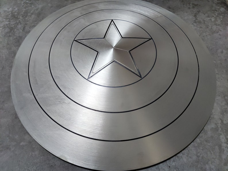 May include: A silver-colored Captain America shield with a five-pointed star in the center. The shield has concentric circles around the star.