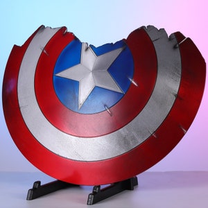 May include: A distressed Captain America shield, featuring a red, white, and blue color scheme with a star emblem at the center. The shield is mounted on a black stand, showcasing a battle-worn appearance with visible damage.