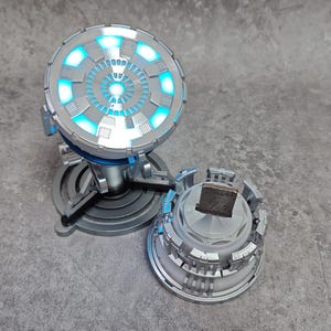 May include: A silver device with a circular design and blue glowing lights, reminiscent of a sci-fi prop. It sits on a stand, with a matching detached component. The design suggests a futuristic or technological theme.