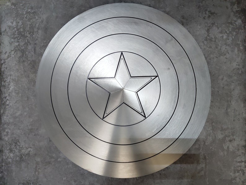 May include: A silver-colored Captain America shield with a red, white, and blue star in the center. The shield has a circular shape with concentric circles around the star.