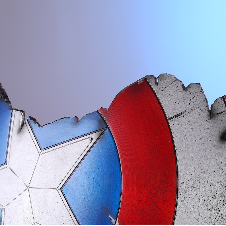 May include: A close-up of a distressed shield with a white star on a blue background, a red stripe, and a silver border. The shield has a battle-worn appearance with chipped edges and a textured surface, suggesting a superhero theme.
