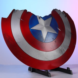 May include: A distressed Captain America shield, featuring a red, white, and blue color scheme with a central star emblem. The shield has a battle-worn appearance, with a large section missing from the top. It is displayed on a black stand.